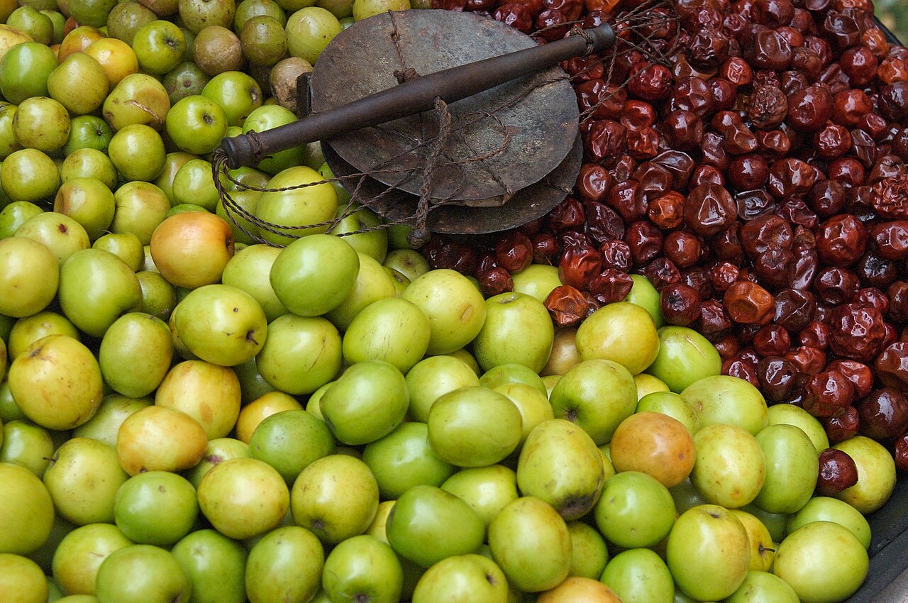 Ber - Green and red boroi ("Ziziphus mauritiana"). I believe the red is just a dried version of the same fruit, in a different colour.
Note the scale, for weighing out larger quantities of fruit.